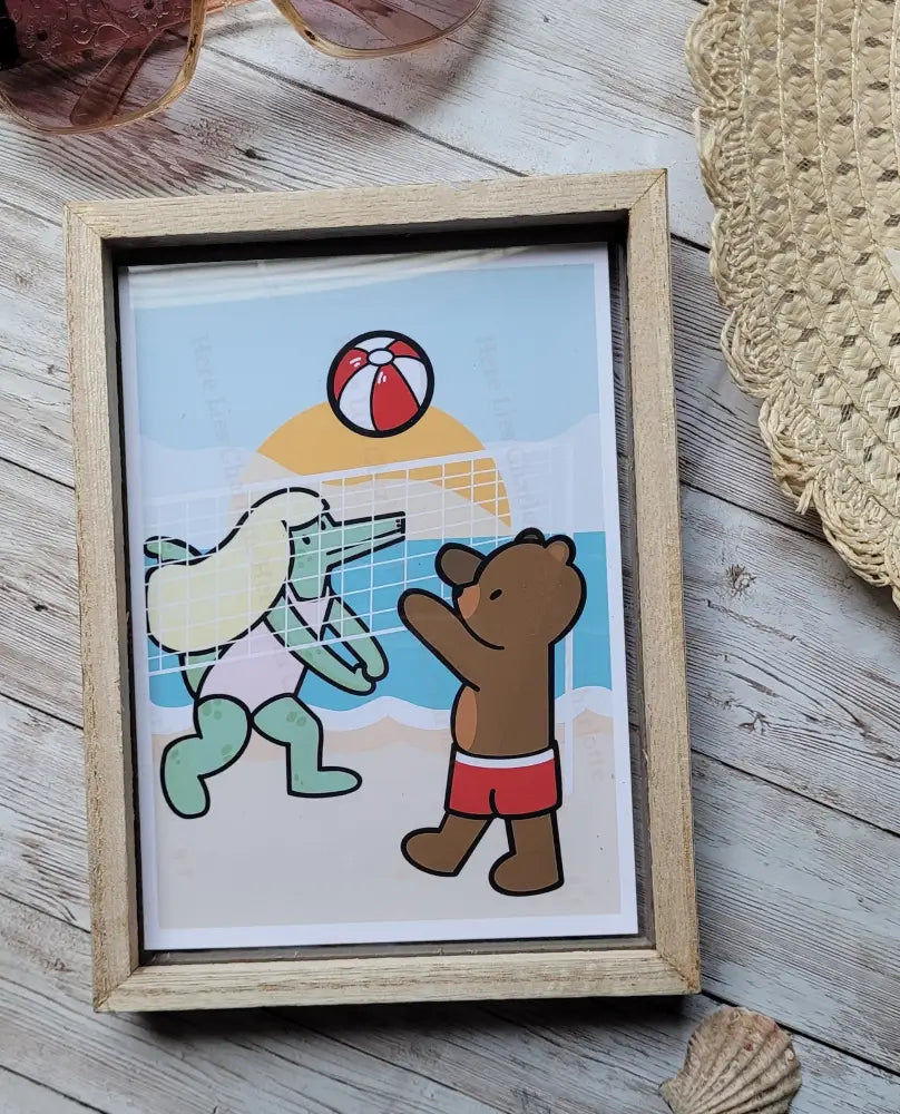 Framed illustration of a bear and an alligator at a beach with a ball, on a wooden surface.