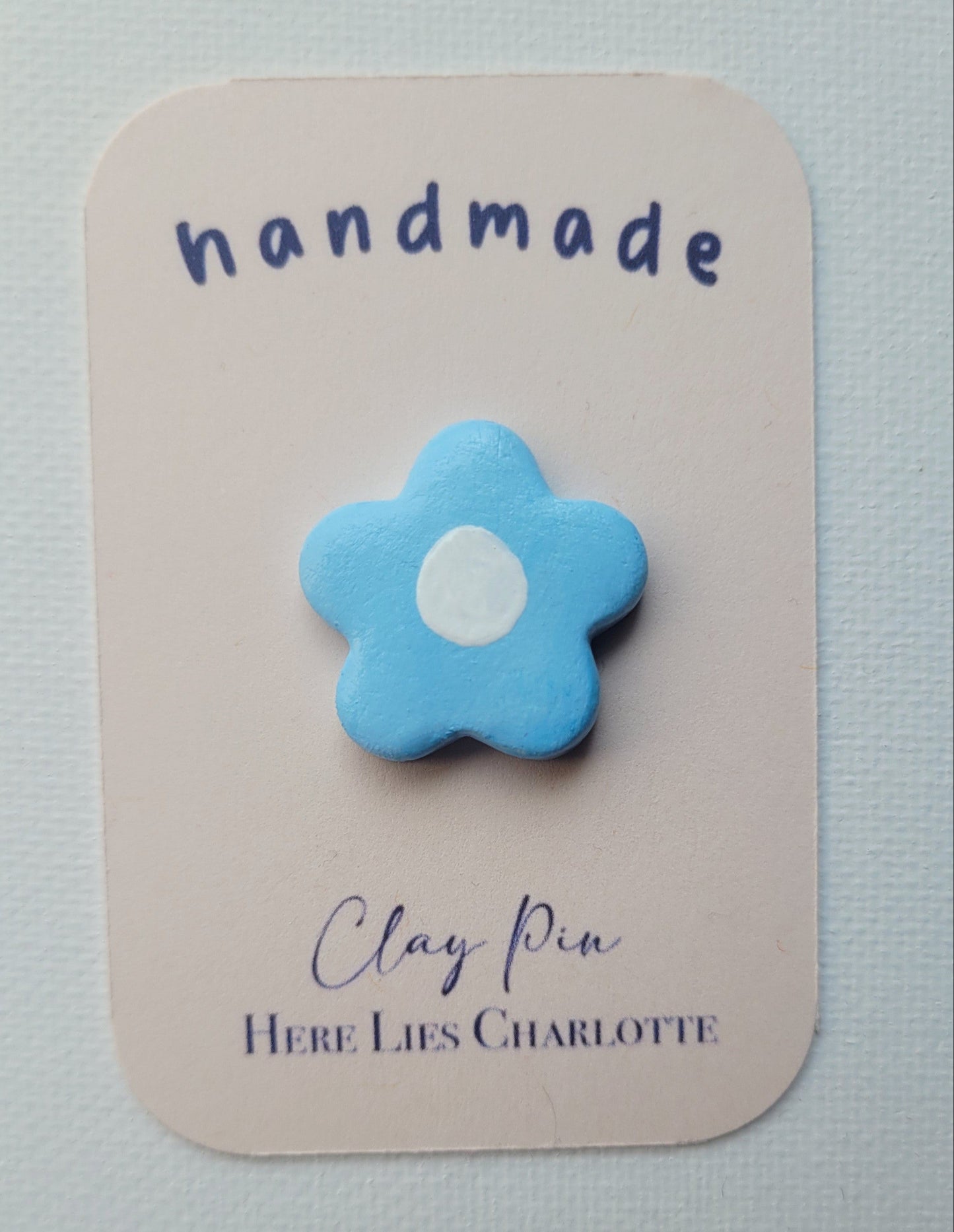 Blue flower-shaped clay pin on a 'handmade' card with 'Here Lies Charlotte' text.