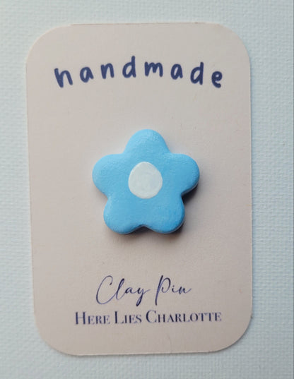 Blue flower-shaped clay pin on a 'handmade' card with 'Here Lies Charlotte' text.