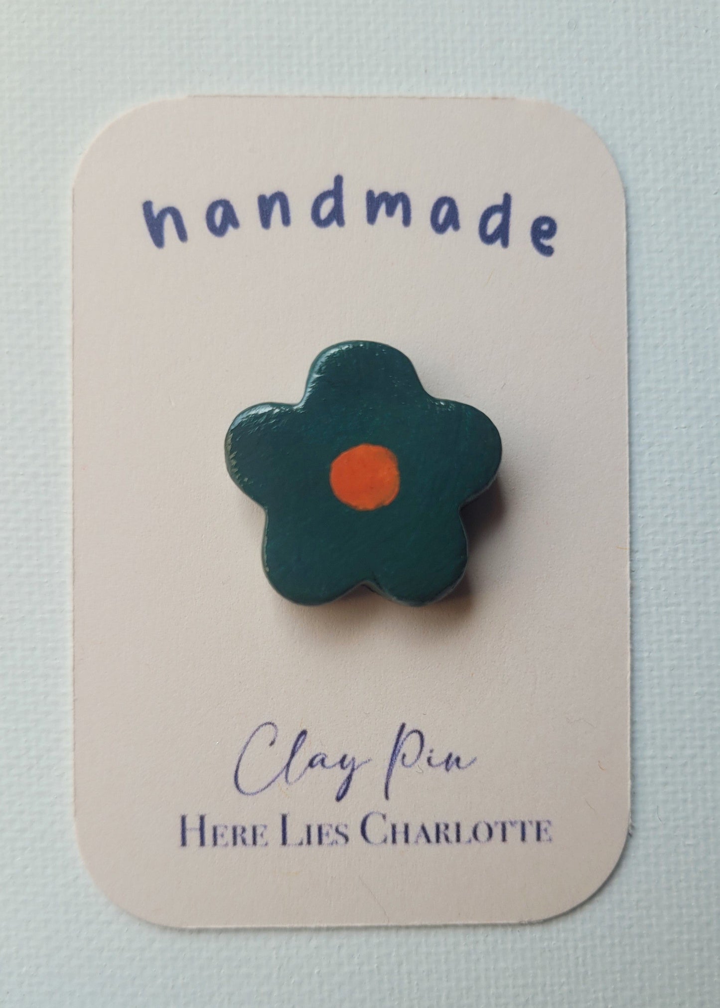 Flower-shaped clay pin on a 'handmade' card with 'Here Lies Charlotte' branding.