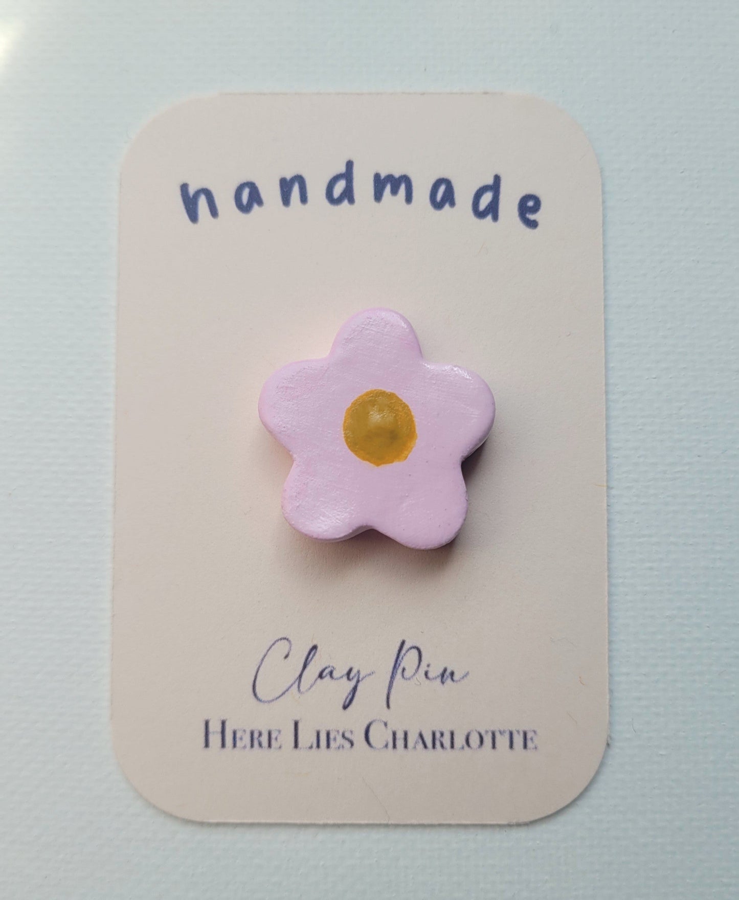 Flower-shaped clay pin on a 'handmade' card with 'Here Lies Charlotte' text.
