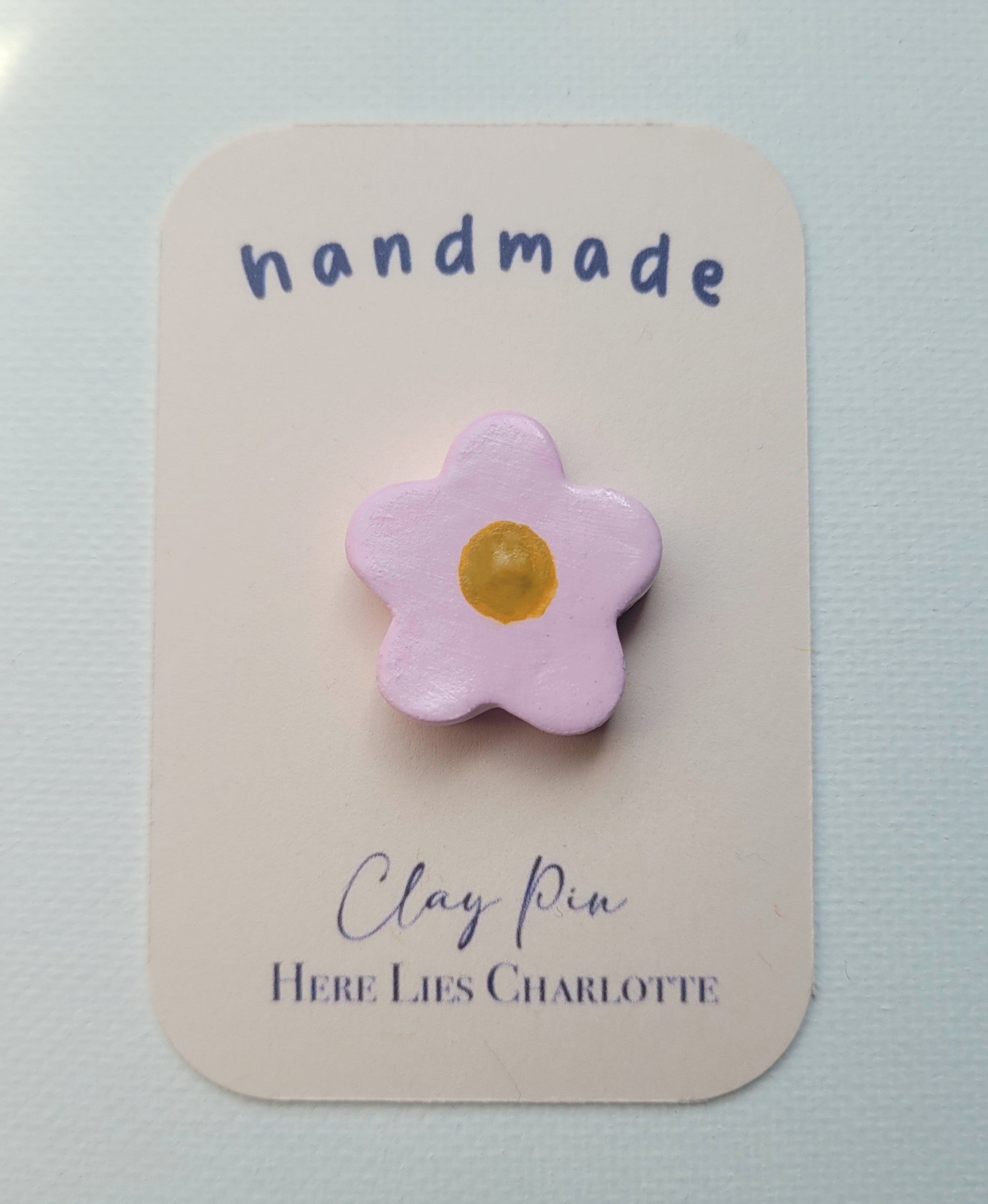 Flower-shaped clay pin on a 'handmade' card with 'Here Lies Charlotte' text.