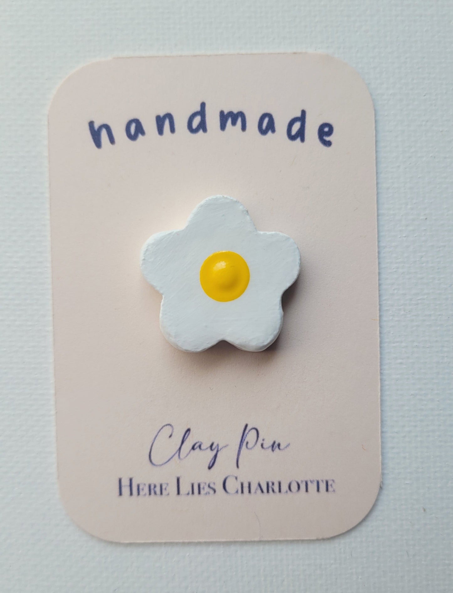 Flower-shaped clay pin on a 'handmade' card with 'Here Lies Charlotte' branding.