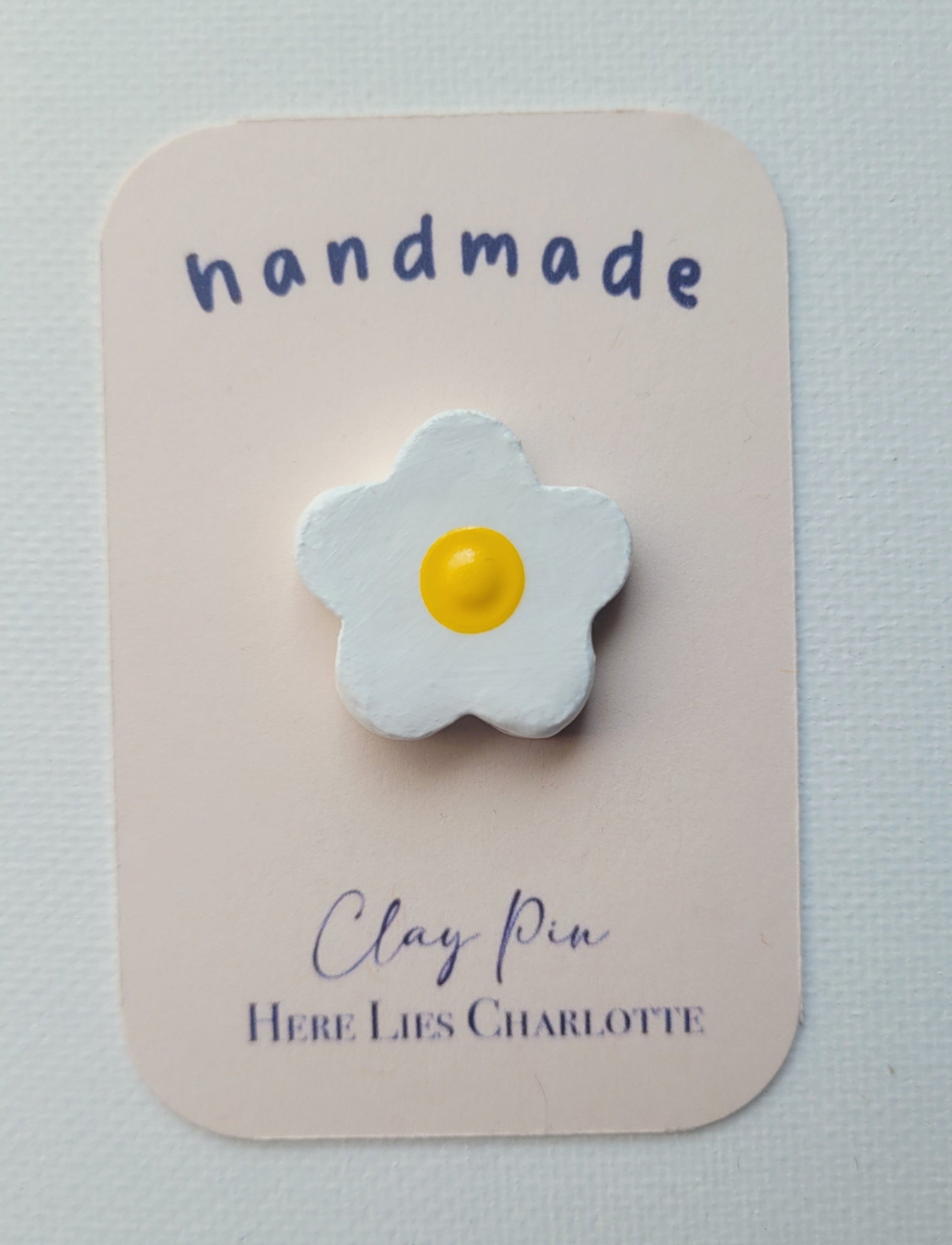 Flower-shaped clay pin on a 'handmade' card with 'Here Lies Charlotte' branding.