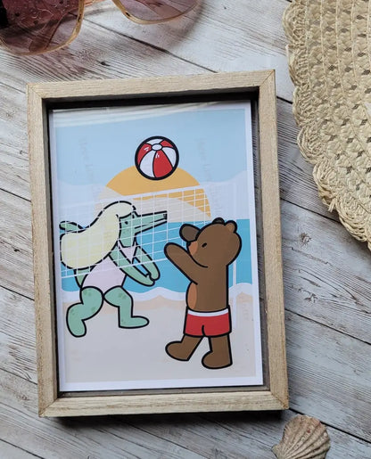 Framed illustration of a bear and an alligator at a beach with a ball, on a wooden surface.